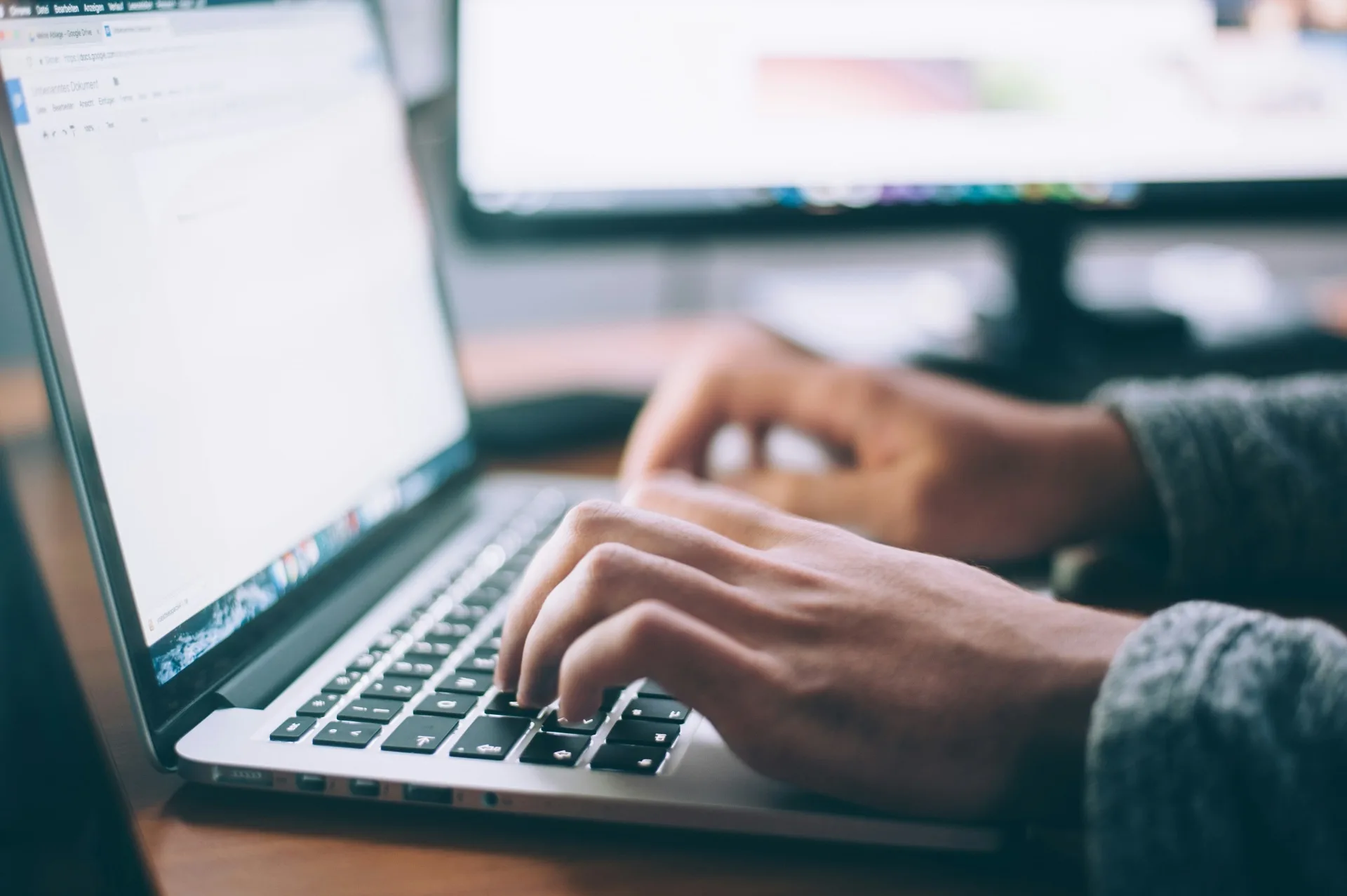 Close-up of hands typing on a laptop keyboard with a second monitor in the background